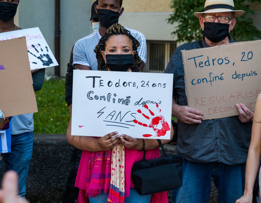 Action menée par Poya Solidaire et Droit de rester. Une femme masquée porte une pancarte sur laquelle on peut lire: Teodore, 24 ans, confiné depuis 4 ans.