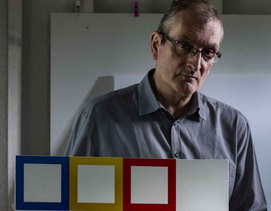 Claudio Bernasconi avec une de ses peintures géométriques, un carré bleu, un carré jaune, un carré rouge, un carré blanc