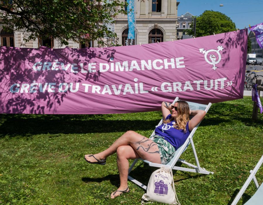 Une femme sur un transat. A l'arrière une banderole violette: "Grève le dimanche, grève du travail gratuit".